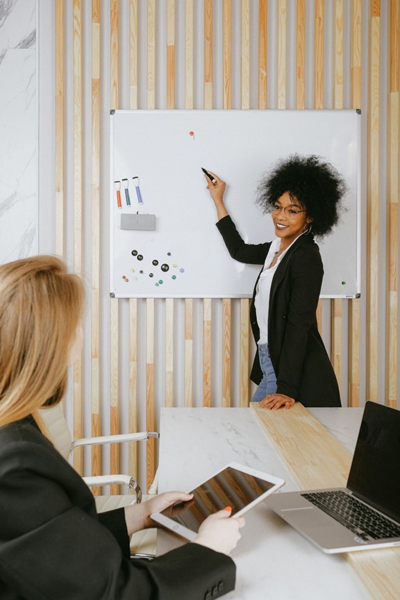 two women, one stands in front of whiteboard coaching the other one that is sitting with tablet in hand