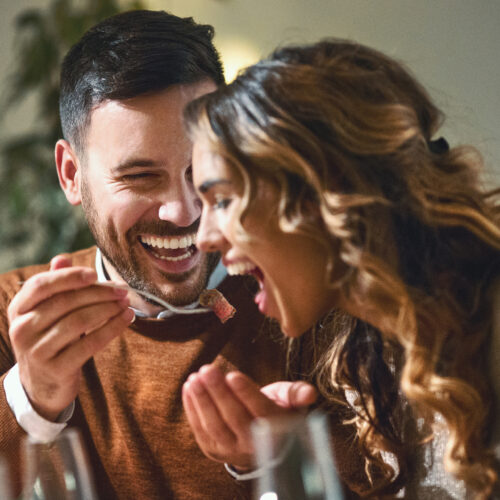 Closeup of mid 20's couple having fun during dinner party. The guy is feeding his girls with some chopped fruit, both laughing. Eating wagyu beef, company using storybrand framework on website and marketing